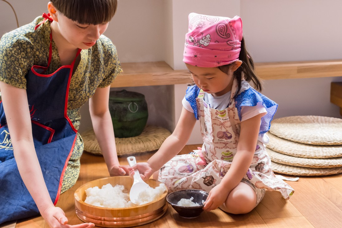 Children in aprons and headbands serving rice from a wooden bowl at Kageoka no Ie community centre.