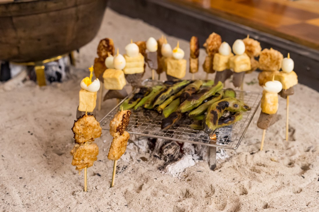 Skewered tsukune chicken meatballs and grilled vegetables cooking on a metal grate over hot coals in a traditional Japanese irori hearth.