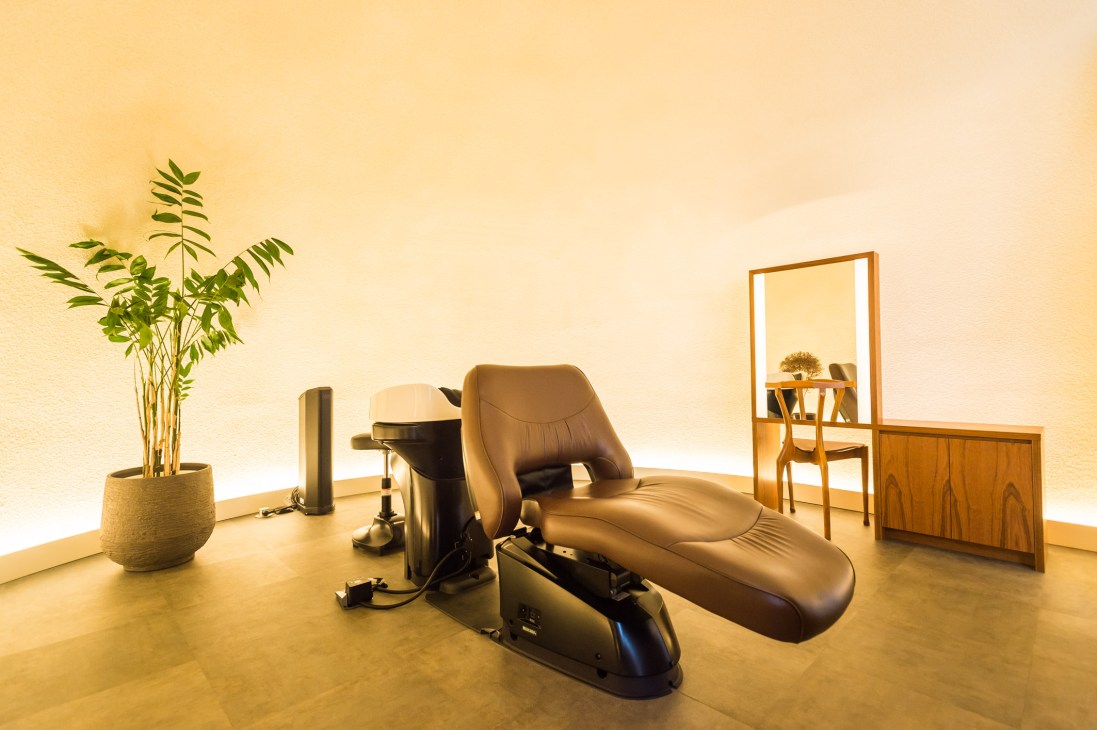 Minimalist spa treatment room with a black leather reclining chair, potted plant, and warm ambient lighting in Linda salon.