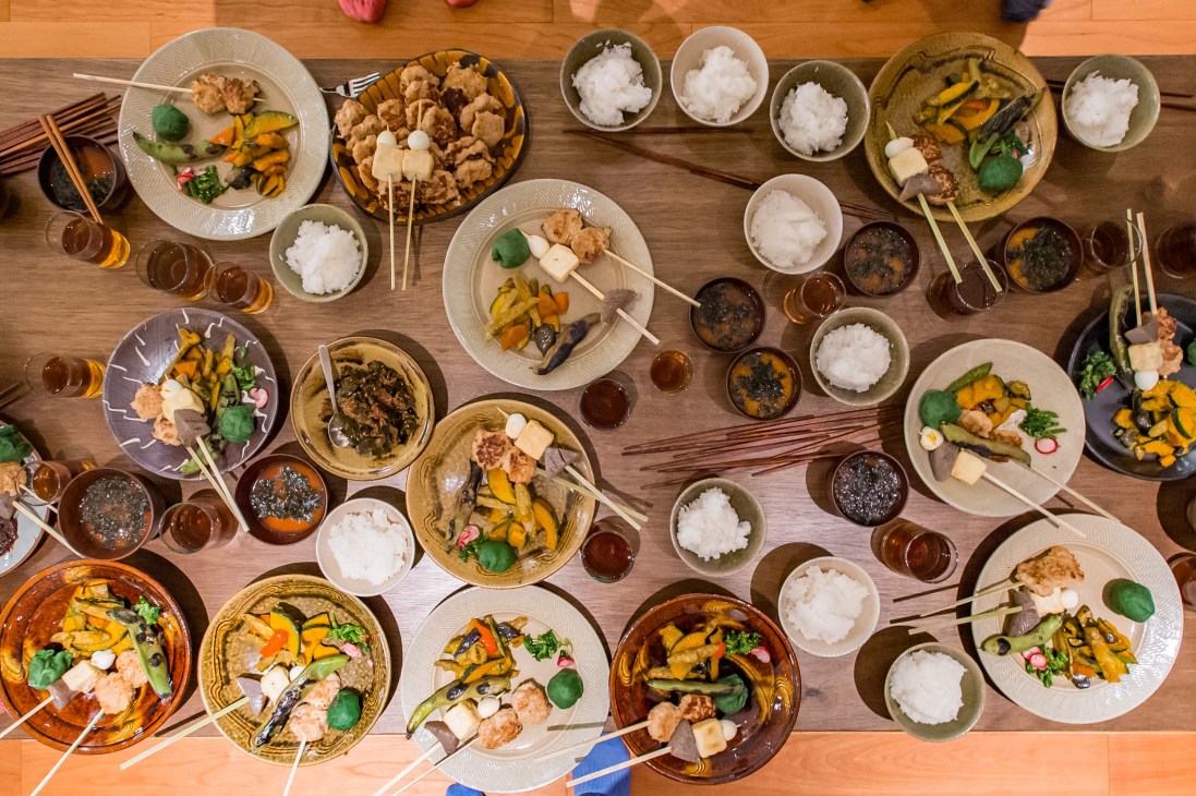 Overhead view of a wooden table with multiple place settings of Japanese food including skewered chicken meatballs, grilled vegetables, and rice bowls.