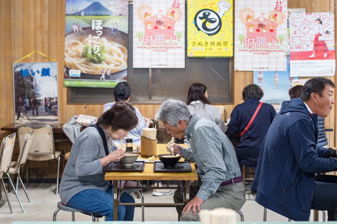 People eating udon noodles at wooden tables in a casual restaurant with Japanese posters on the walls