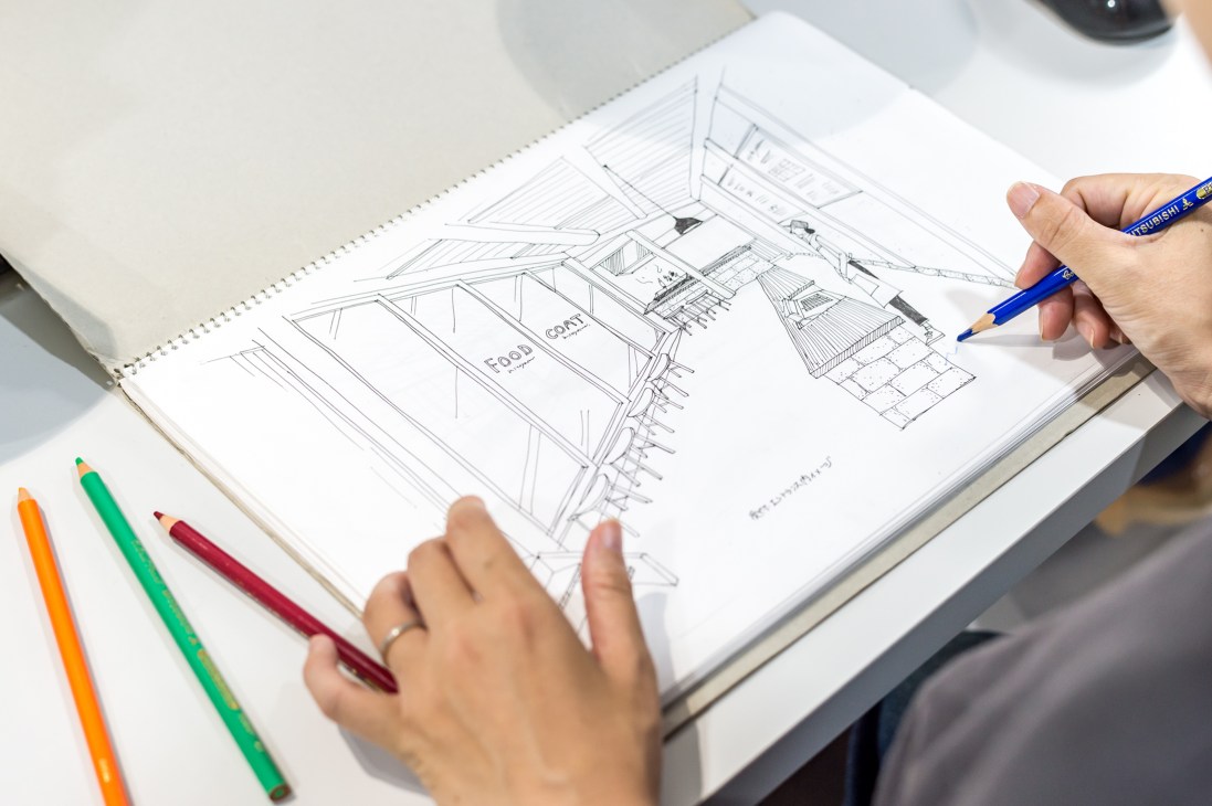 Designer sketching architectural drawings with colored pencils on spiral notebook.