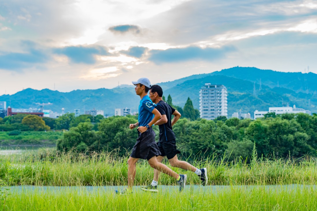 Two men jogging through green fields by a river with mountains and city buildings in the background.