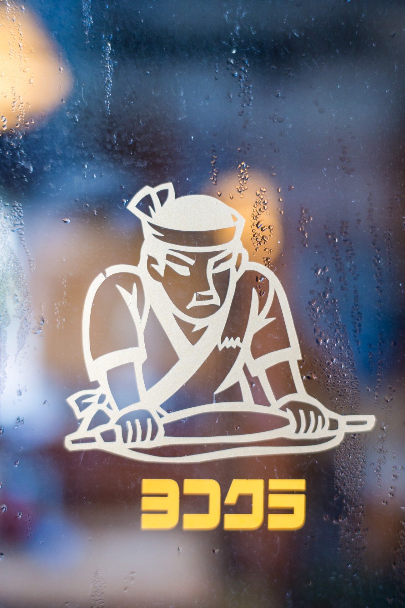 White stylized figure of a person making noodles with rolling pin on rain-covered window with Japanese characters below