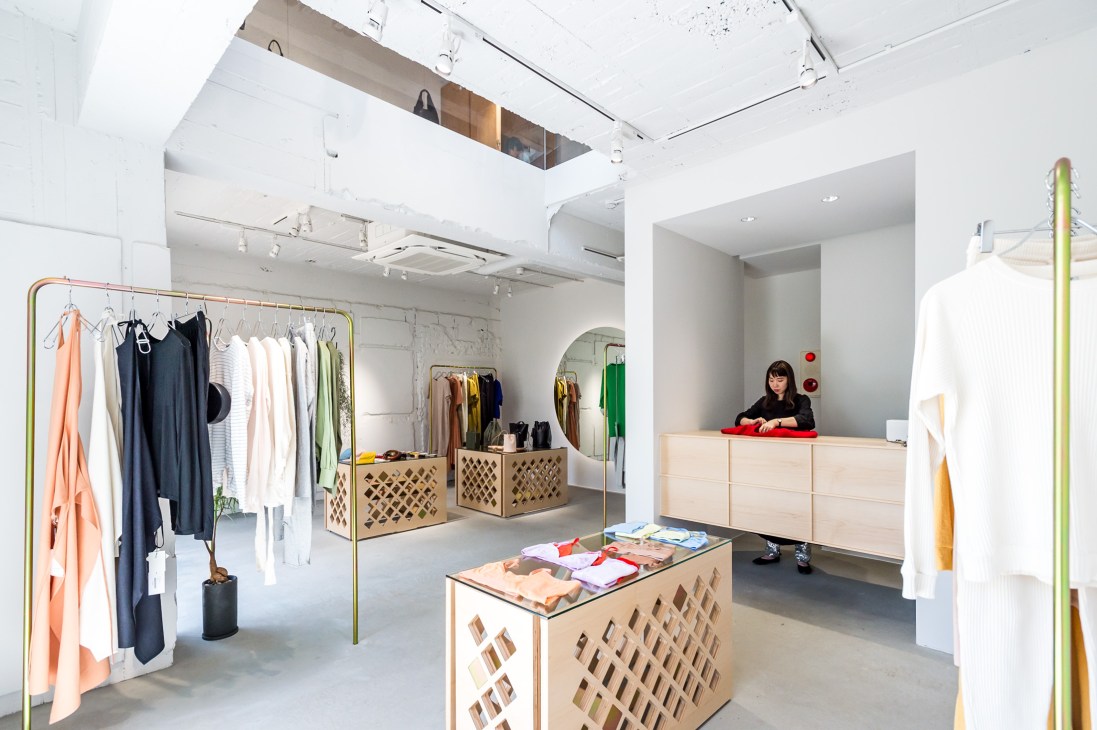 Bright minimalist fashion retail interior with mobile clothing racks, wooden display units, and a staff member at the counter.