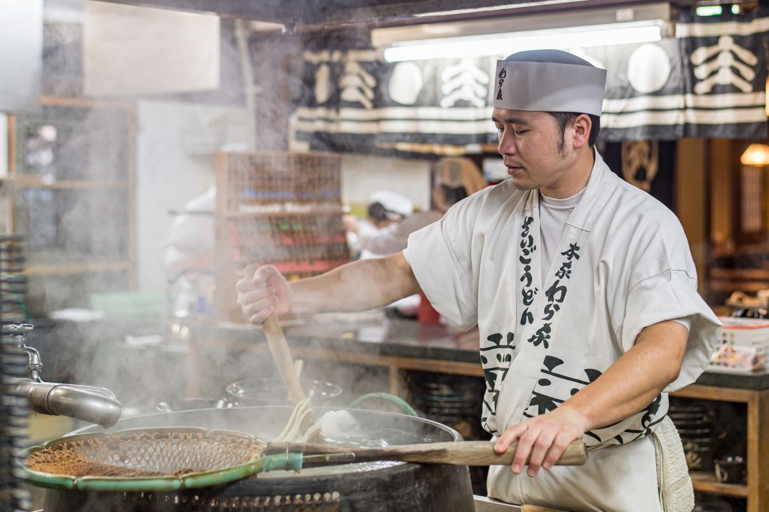 Chef in traditional white uniform and headband cooking noodles in steamy kitchen at Waraya restaurant