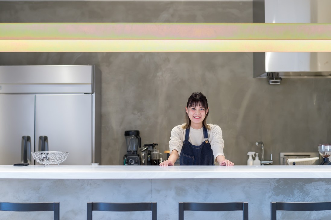 A smiling barista stands behind the counter of a modern café with minimalist design and concrete walls.