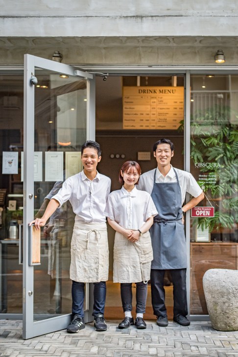 Three smiling staff members in white shirts and aprons stand in the doorway of Obscura Coffee Roasters in Hiroshima.