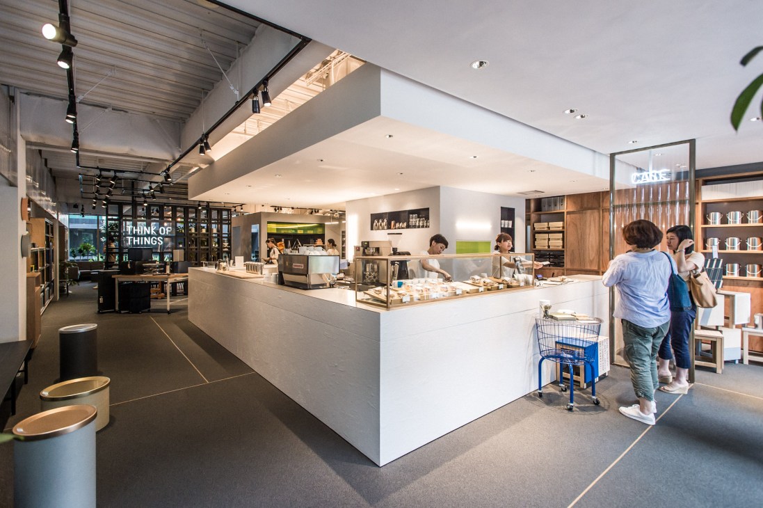 Modern cafe counter with white surfaces, pastries in glass case, customers ordering, and exposed ceiling