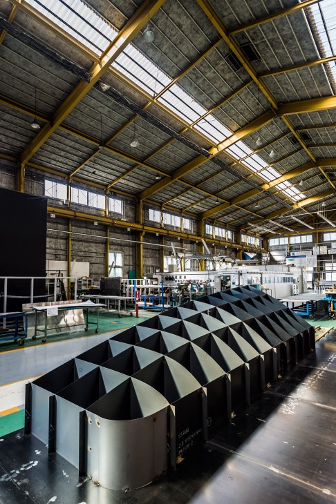 Industrial factory interior with yellow steel beams, metal workstations, and geometric black and white metal components on the floor.