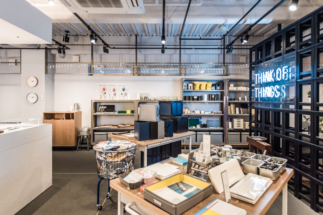 Modern stationery store interior with wooden display tables, industrial shelving, and blue neon sign reading 'THINK OF THINGS'