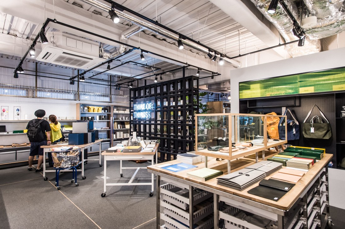 Modern retail interior with white tables displaying stationery and notebooks, black shelving units, and customers browsing