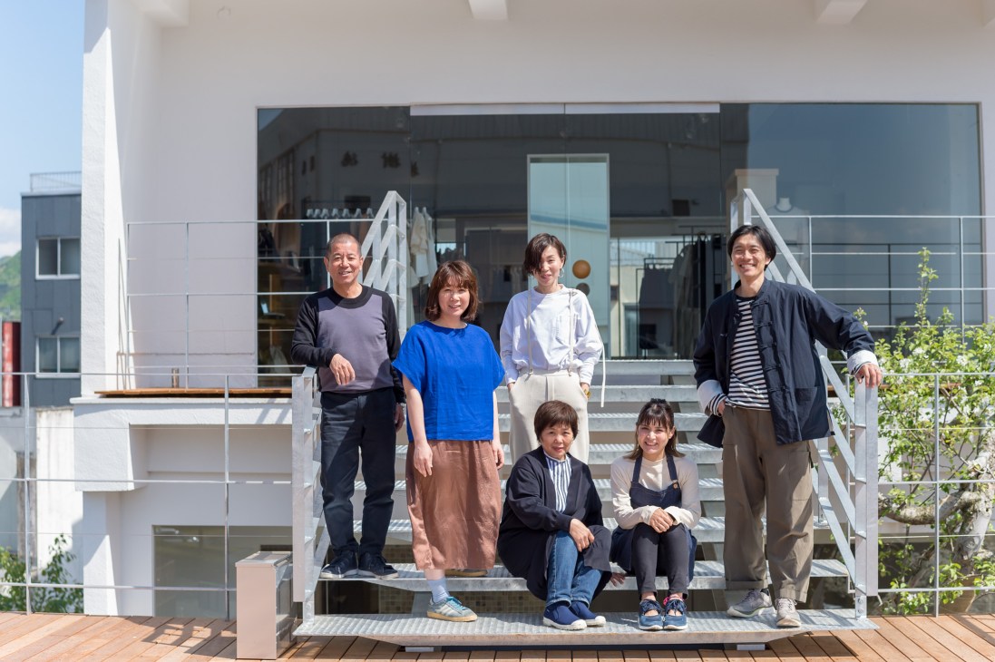 Toshio Tamaki and his team of seven people posing on a wooden terrace outside ST Company fashion shop.