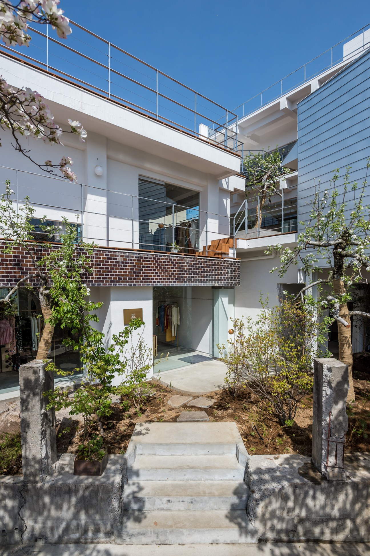 Modern multi-level fashion shop with white and grey exterior, blooming trees, and metal railings in Kiryu, Japan.