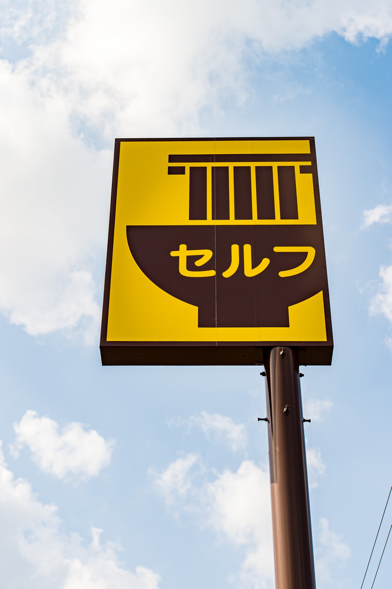 Yellow sign with Japanese characters セルフ and an illustration of a noodle bowl against cloudy sky