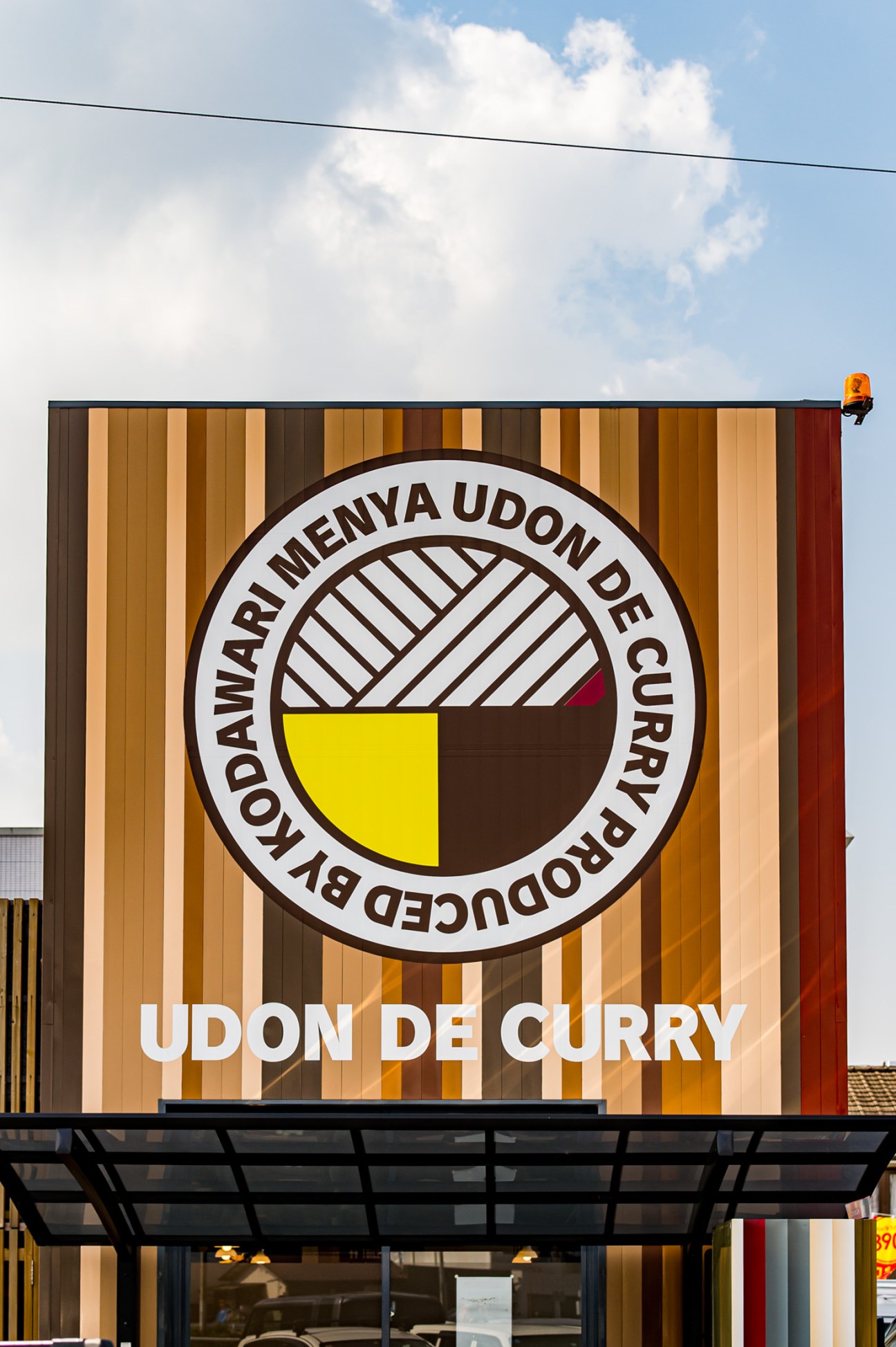 Storefront of Udon De Curry restaurant with brown striped exterior and circular logo