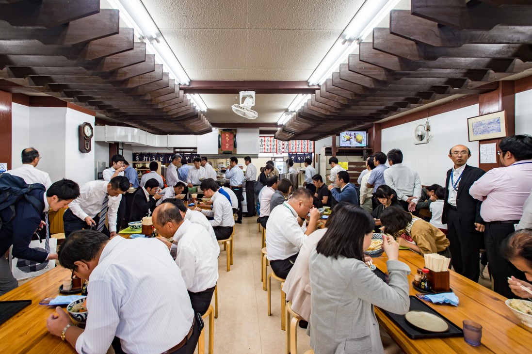 Crowded interior of Sakaeda udon restaurant in Takamatsu with diners at wooden tables and staff working