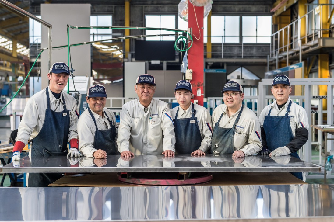 Six Kikukawa metalworkers wearing company caps and aprons pose together at their factory workstation.