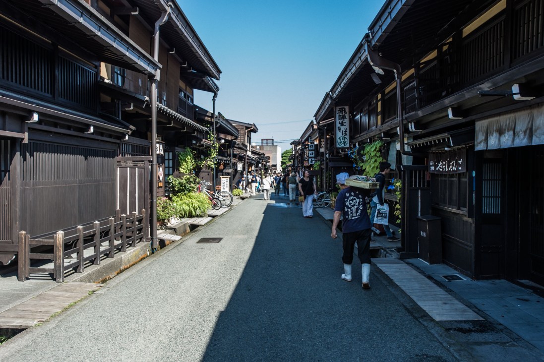 A narrow pedestrian street in Takayama lined with traditional dark wooden buildings and shops.