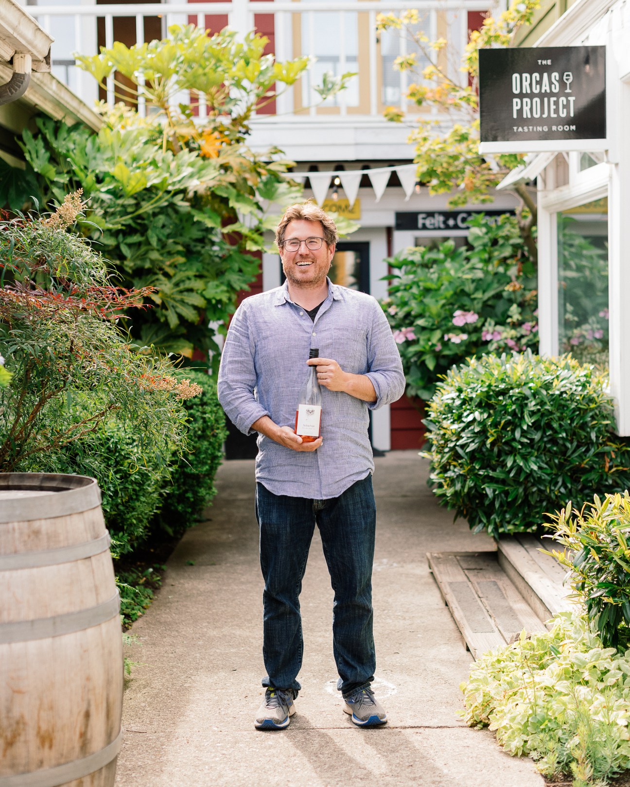 Cole Sisson holding wine bottle outside Orcas Project Tasting Room surrounded by lush garden plants