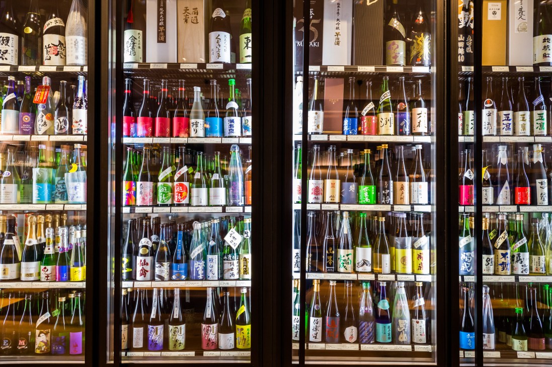 Display cases filled with colorful bottles of Japanese saké, shochu, and other spirits arranged on illuminated shelves.