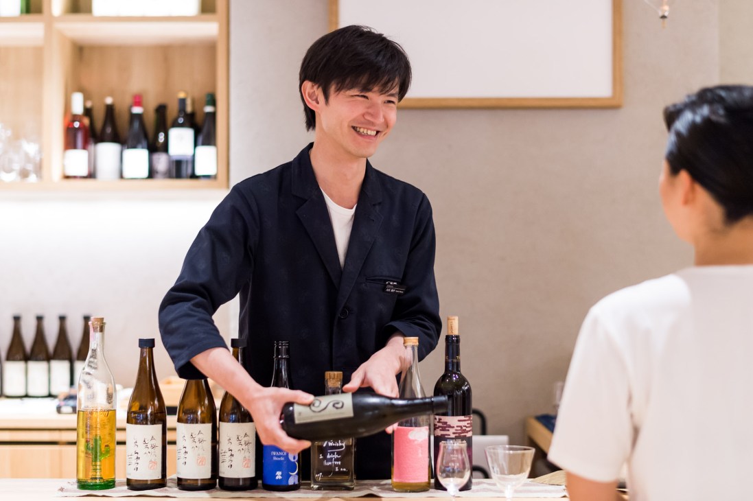 Shop manager Shohei Okawa pouring a bottle at Imadeya's tasting counter with various sake and liquor bottles displayed.