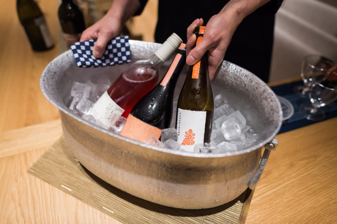 Hands holding wine bottles in an ice bucket during a tasting session at Imadeya liquor shop.