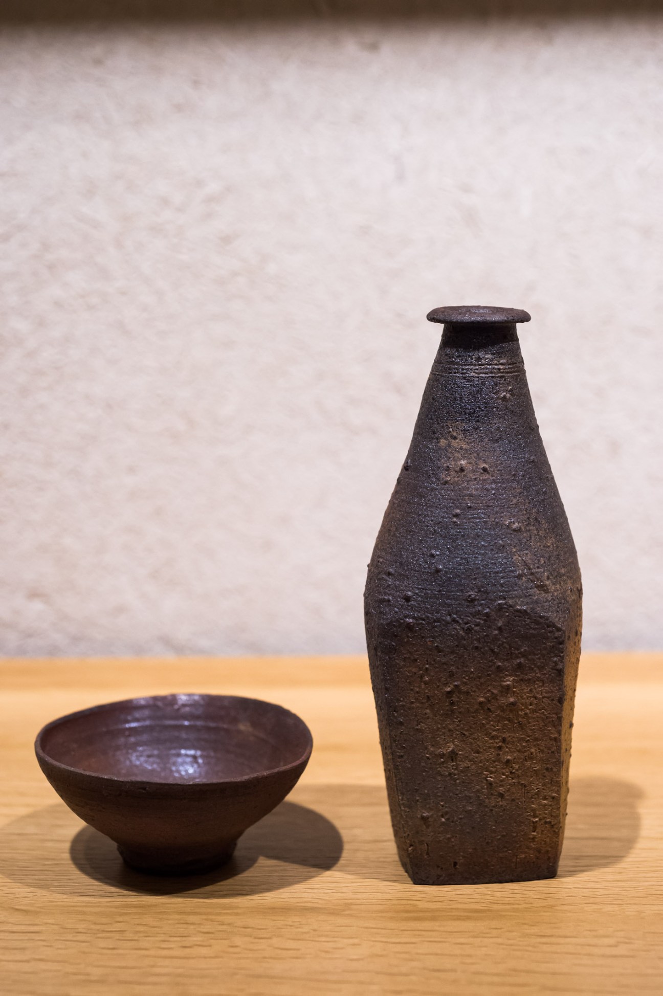 Dark ceramic bottle and small bowl displayed on wooden surface against white wall.