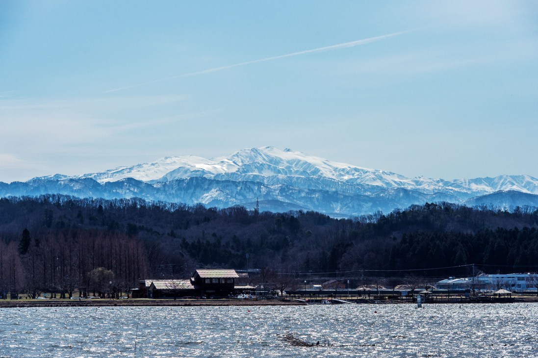 Snow-capped Mount Haku rises beyond forested foothills and a rural lakeside settlement in Japan's Hokuriku region.