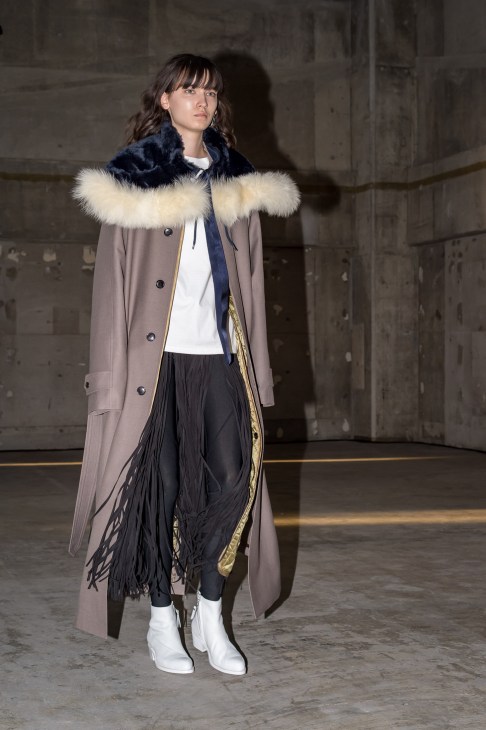 Model wearing a taupe coat with fur collar, black pleated skirt and white boots on a runway.