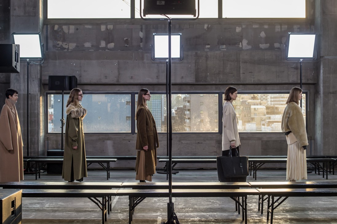 Models in neutral-toned coats rehearse on a runway in front of large industrial windows overlooking a city skyline.