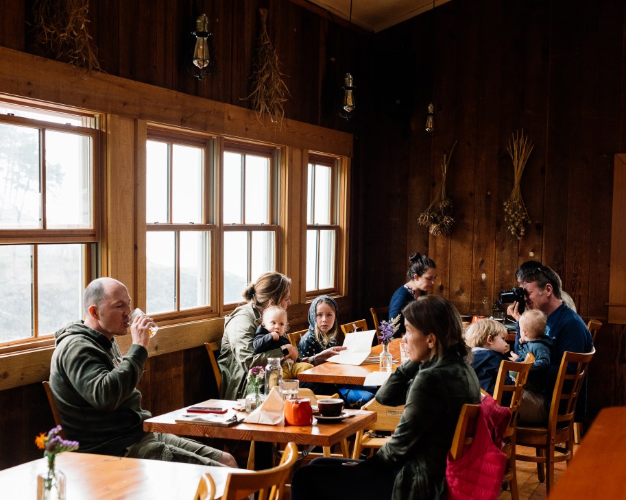 Families dining at wooden tables in rustic restaurant interior with large windows and exposed beams