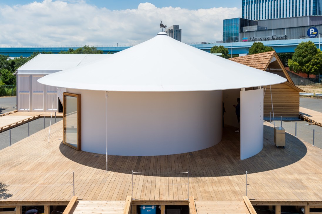 Hiragana-no Spiral House with white conical roof and curved walls on wooden deck beside water.