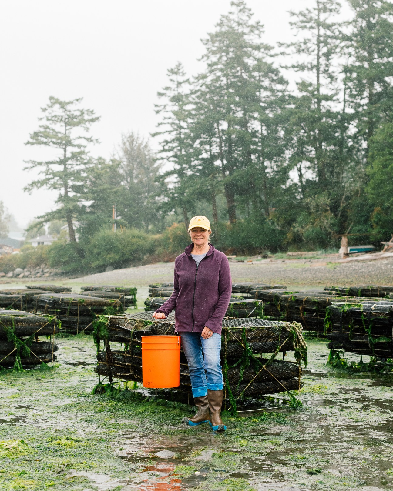 Toni Knudson stands smiling among shellfish traps with an orange bucket at Buck Bay Shellfish Farm