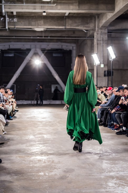 Model wearing a green flowing dress with black belt on a runway at Hyke fashion show.
