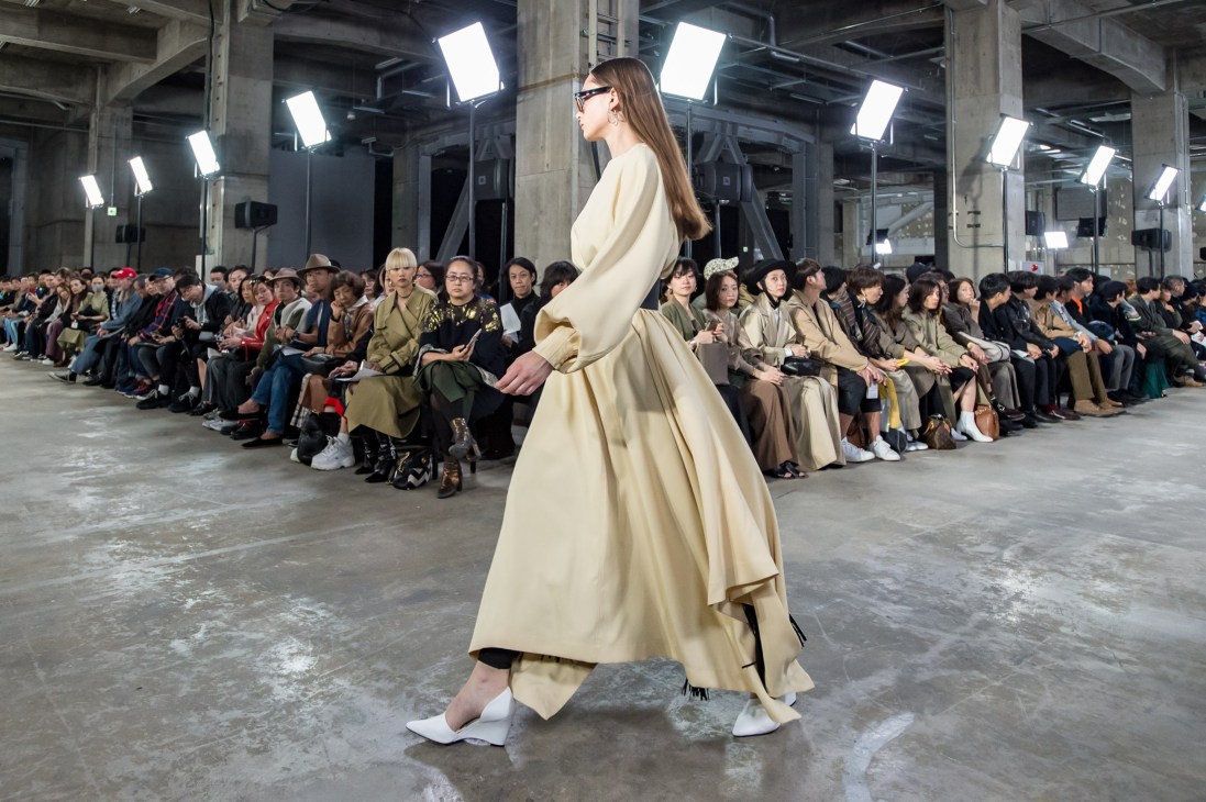 Model in cream-coloured flowing coat walking on warehouse runway at Hyke fashion show with seated audience.