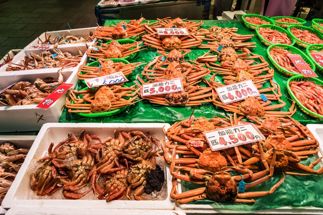Fresh crabs displayed on green plastic trays and white containers at a market stall with Japanese price tags.