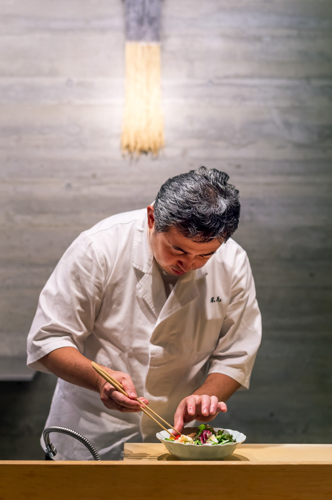 Chef Nobuki Watanabe in white chef's coat preparing a salad with chopsticks at Yoneka restaurant in Tokyo.