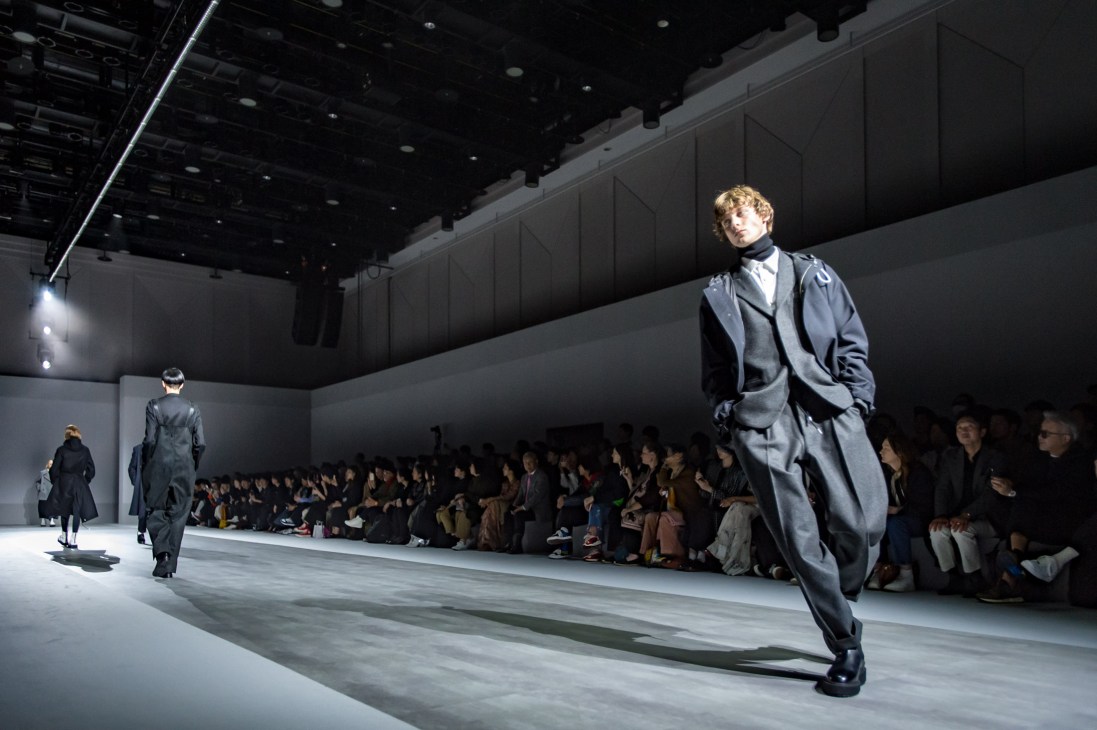 Models walking on a runway during a fashion show with audience seated along the sides in a large venue.