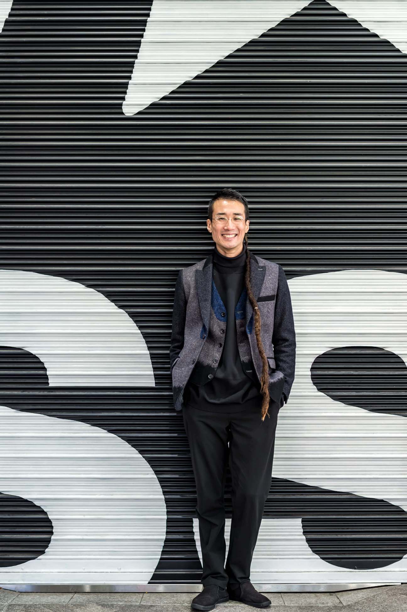 TeamLab's Takashi Kudo stands smiling in front of a black and white striped wall with geometric shapes.