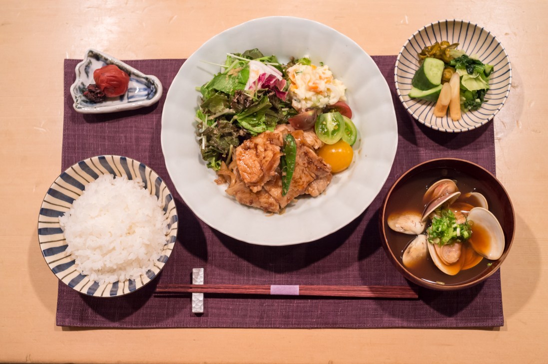 A Japanese meal set featuring ginger pork, rice, miso soup with clams, pickled vegetables, and garnishes on a purple placemat.