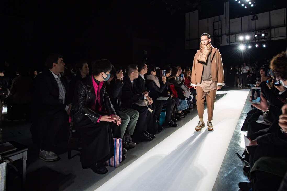 Model walking down a lit runway at Tokyo Fashion Week wearing a beige outfit and headscarf, with seated audience members and photographers on both sides.
