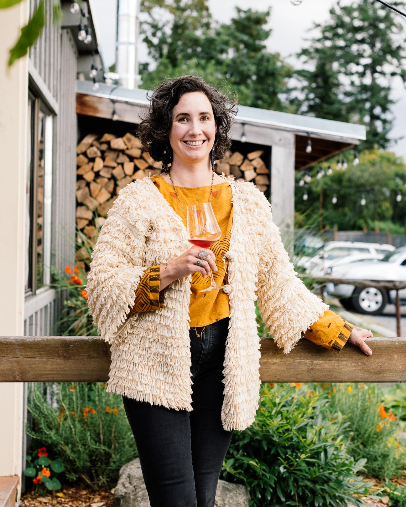 Woman with curly hair in cream textured jacket and yellow top holding wine glass outdoors