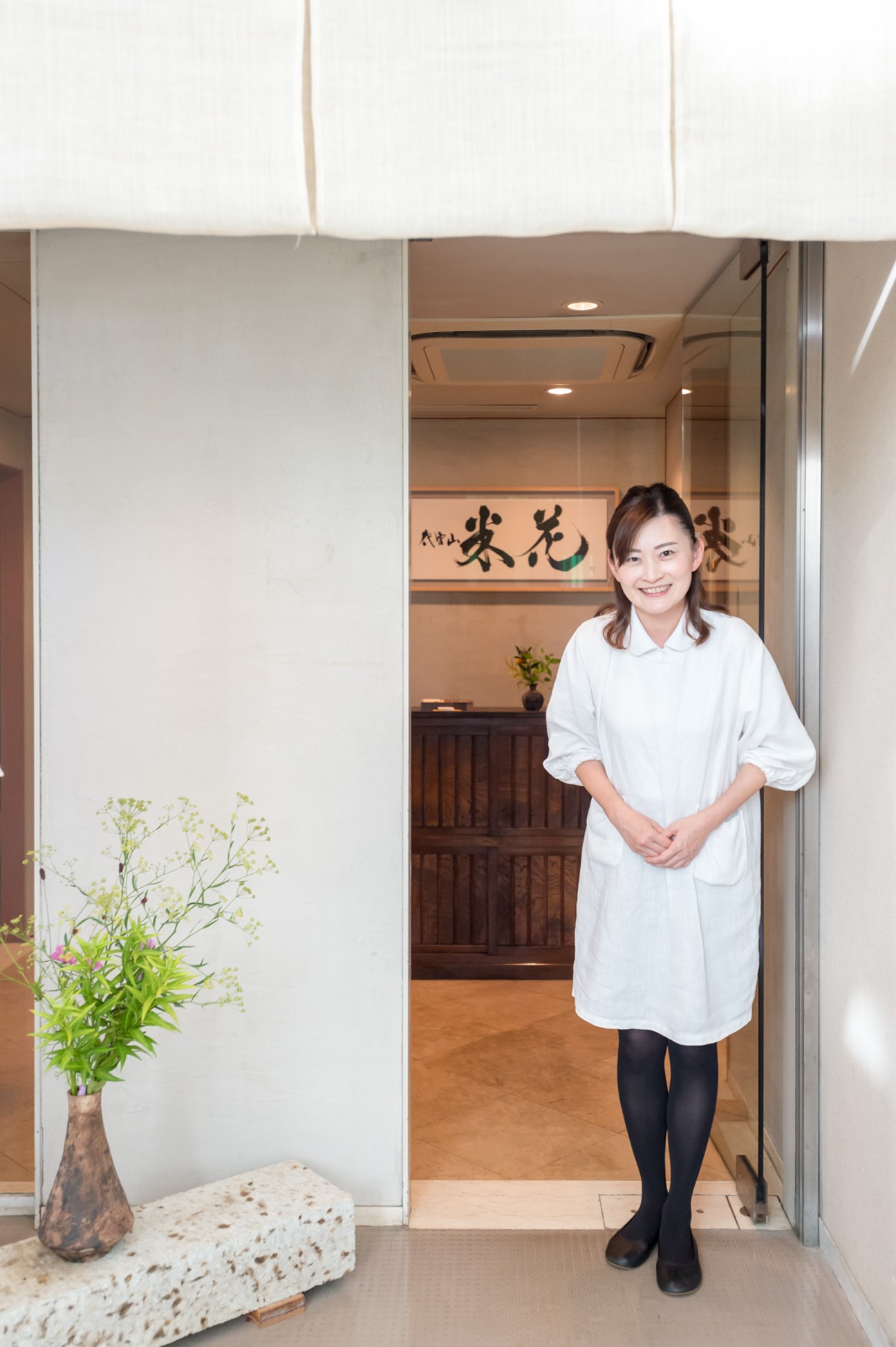A woman in a white shirt stands smiling at the entrance of Yoneka restaurant, with Japanese calligraphy visible on the wall behind her.