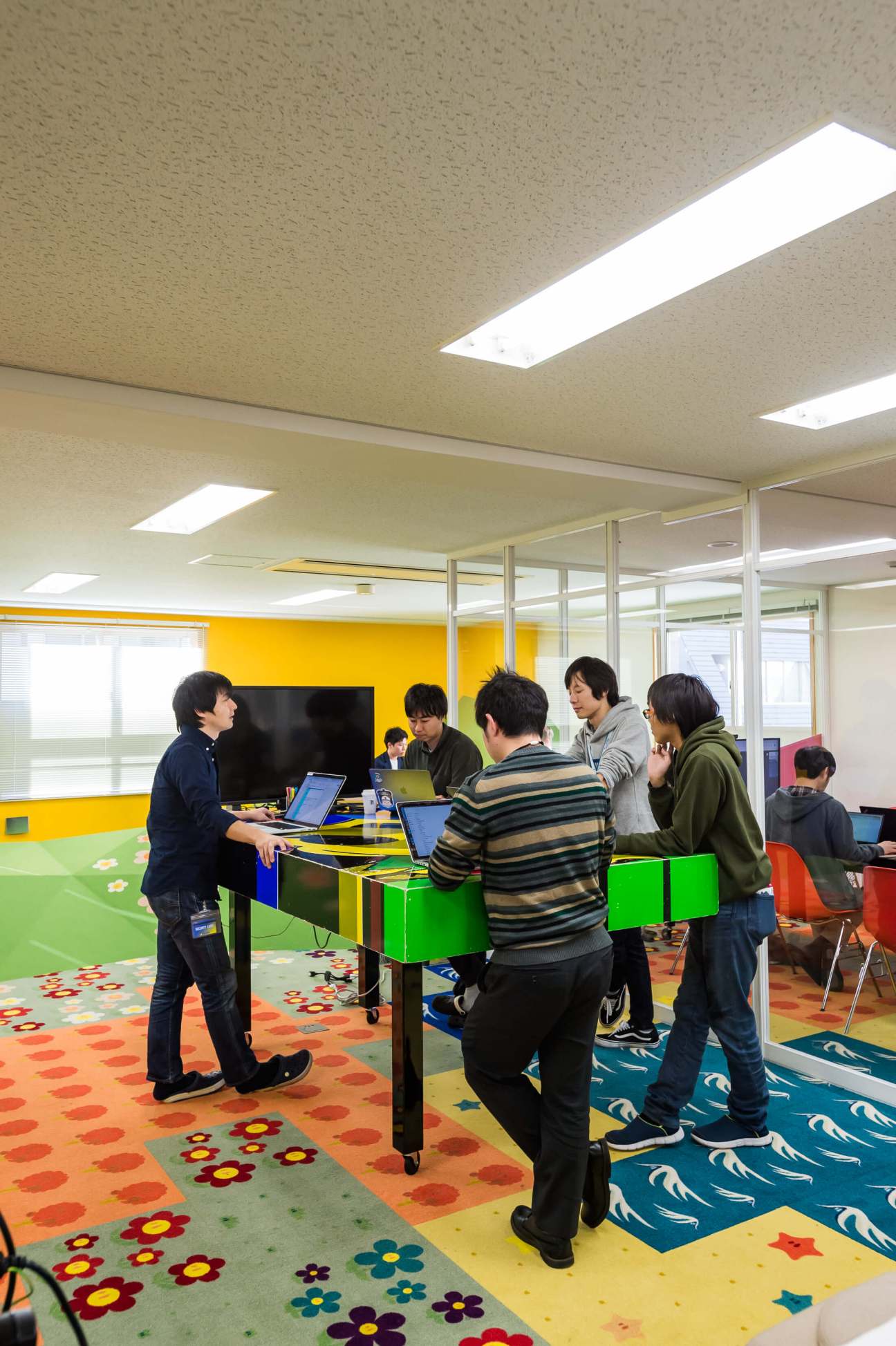 TeamLab employees collaborating around a colorful table with laptops in a bright office space with patterned flooring.