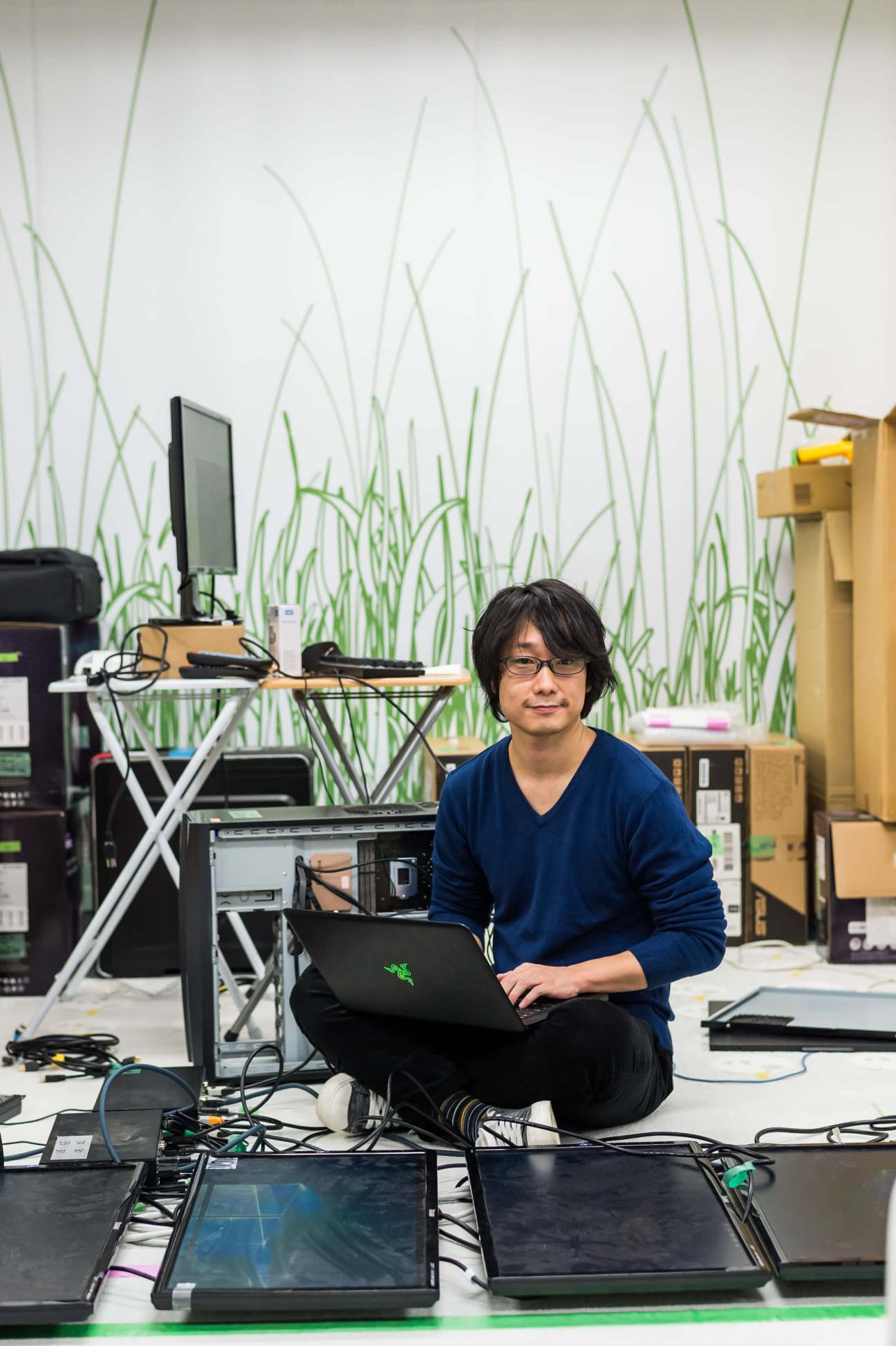 Software engineer Hidekazu Fukunaga sits with a laptop in TeamLab's Tokyo office, with a grass-patterned wall behind him.