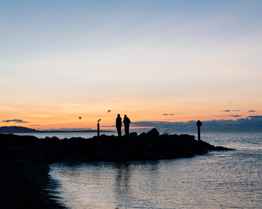 Two silhouetted figures standing on rocky jetty at sunset overlooking water with birds flying overhead