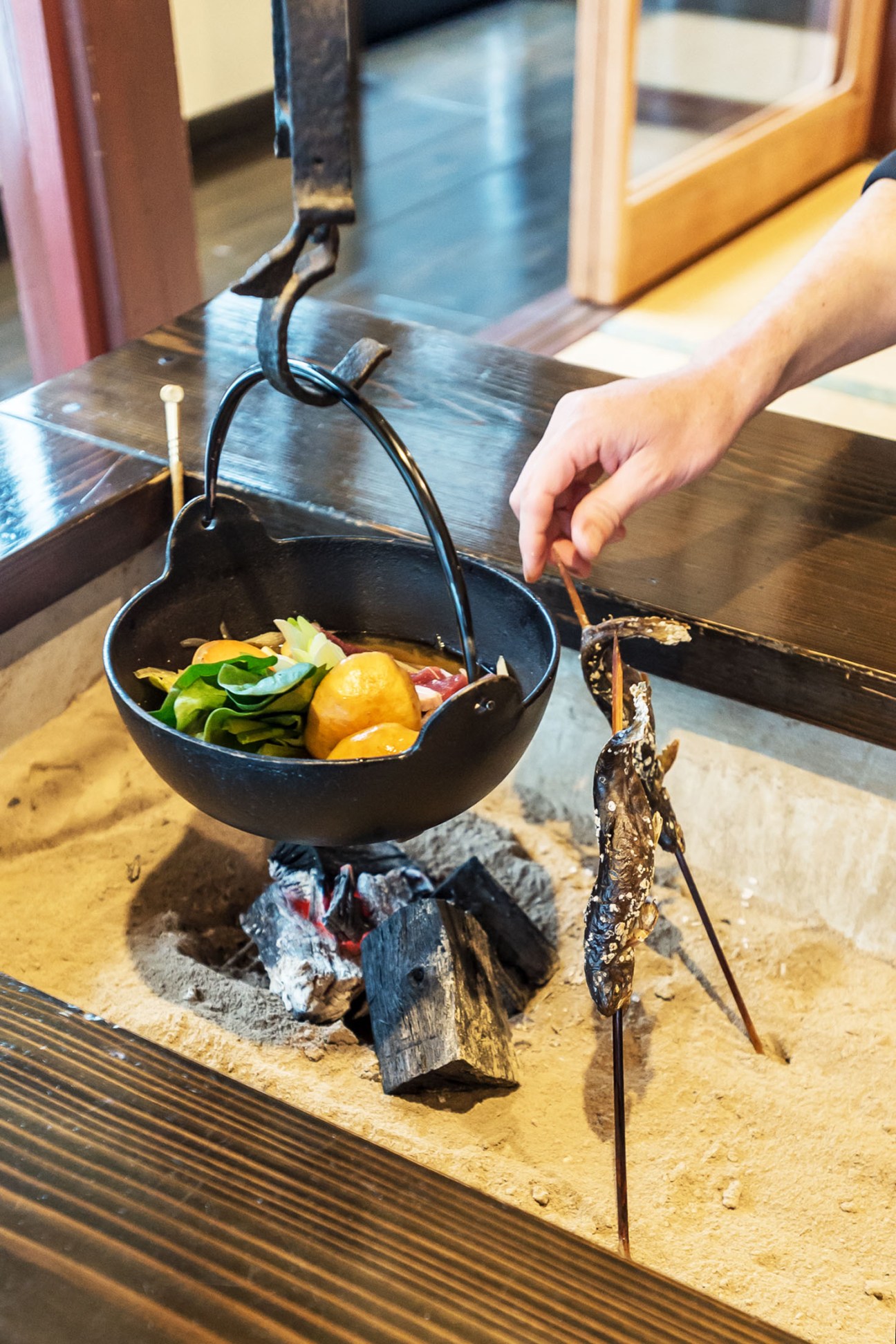 Hand holding chopsticks over a black cast-iron pot of boar hotpot suspended over a charcoal fire at Furari.