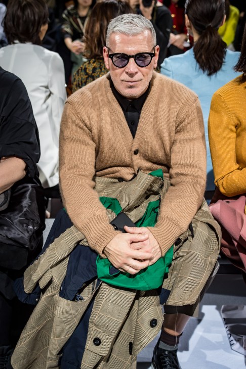 Fashion consultant Nick Wooster seated at Tokyo Fashion Week wearing sunglasses, camel cardigan and patterned trousers.