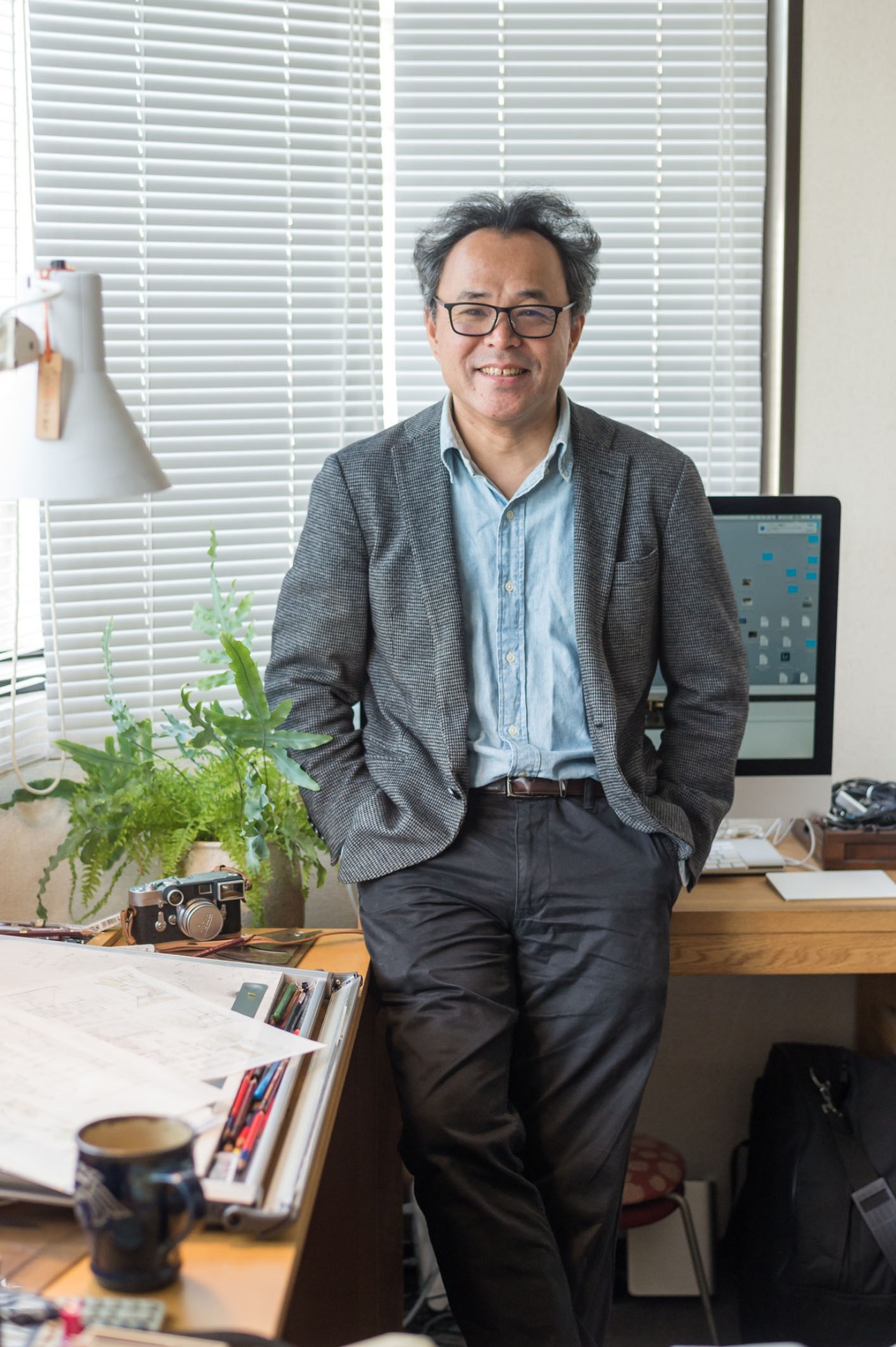 Architect Yasushi Horibe stands in his office smiling at the camera, with design materials and a camera on his desk.